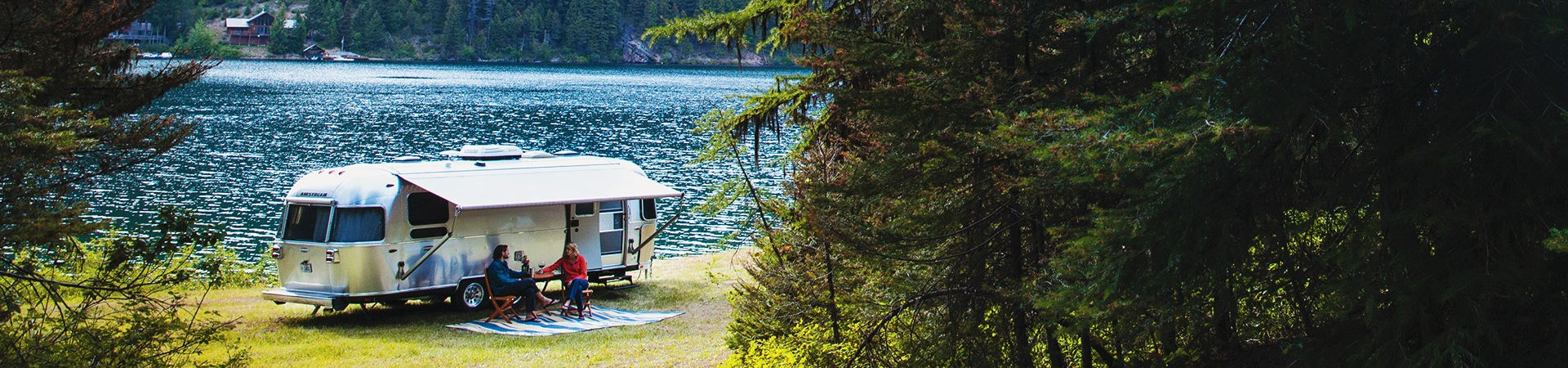 Couple having wine next to their Airstream travel trailer next to a wooded lake